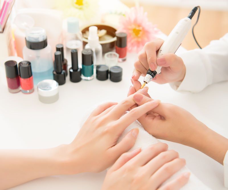 Professionelle Maniküre: Die Nägel einer Kundin werden mit einem Nagelfräser bearbeitet, im Hintergrund Nagellacke